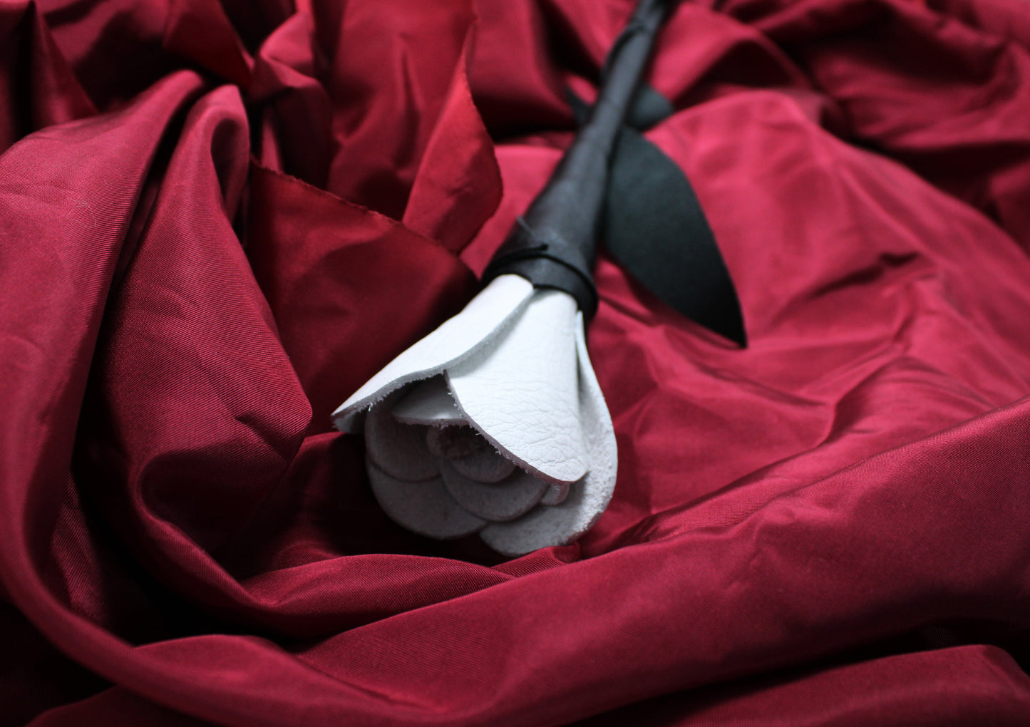 rose bdsm en cuir blanc, avec détail sur la fleur ouverte, et tige en cuir noir, sur fond de satin bordeaux
